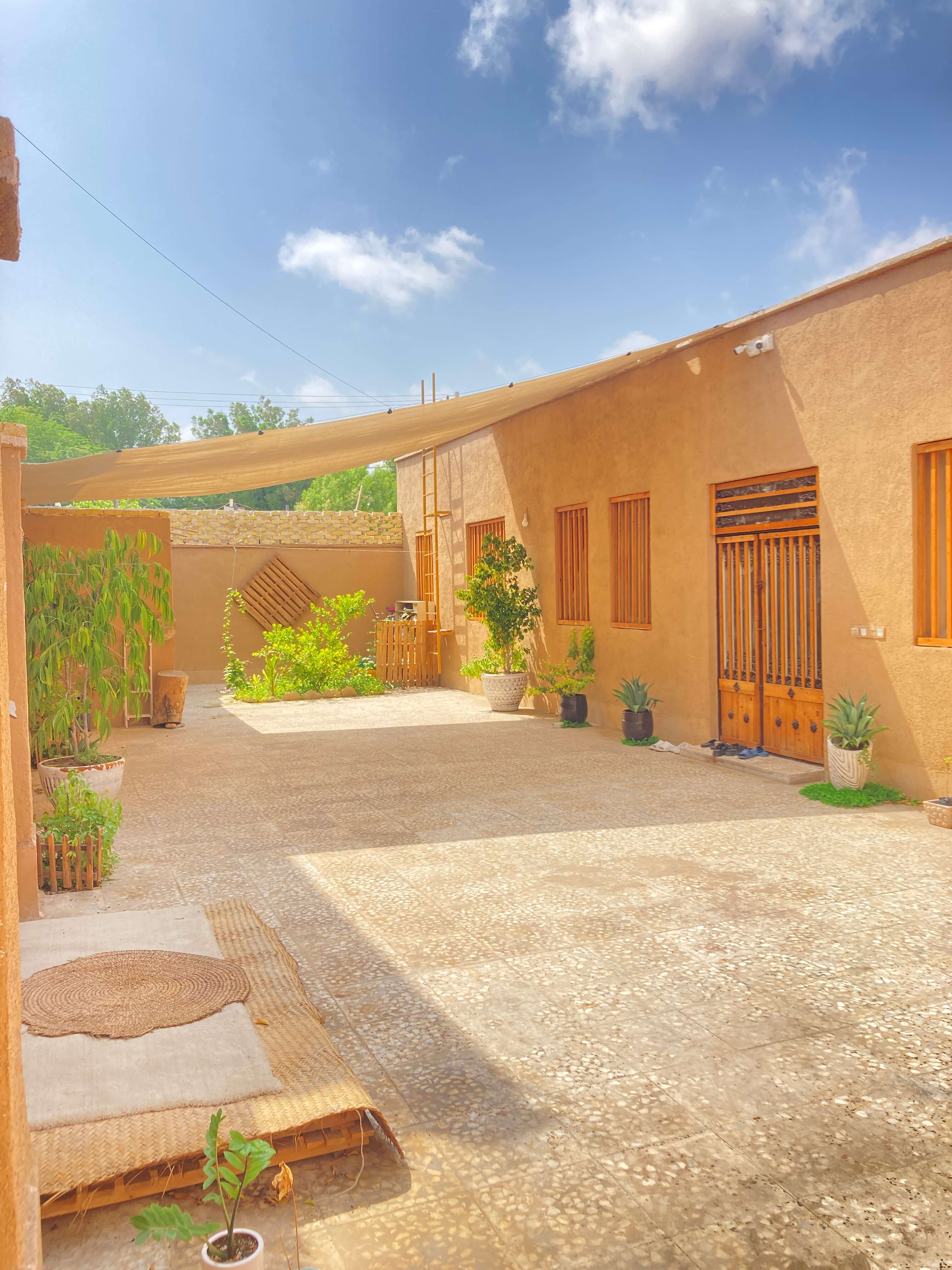 The sunlit courtyard of Bird Hostel in Bandar Abbas, with adobe walls, wooden doors and green plants under a canvas shade